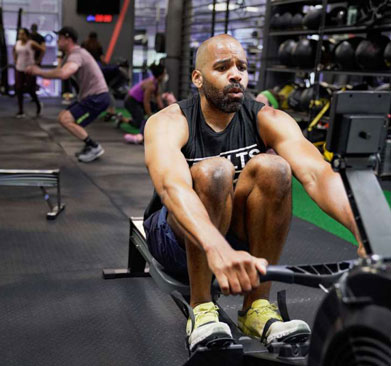 Man performing a rowing exercise on an indoor rowing machine during a coached group fitness workout at a gym, with other members training in the background